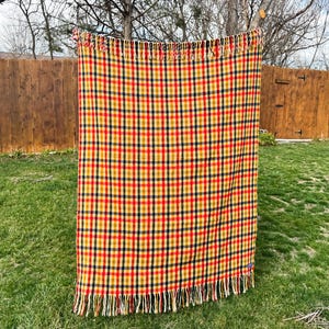 May include: A rectangular throw blanket featuring a vibrant plaid pattern in shades of yellow, red, and dark blue. The fabric has a woven texture and is finished with long, cream-colored fringe along the top and bottom edges of the textile.