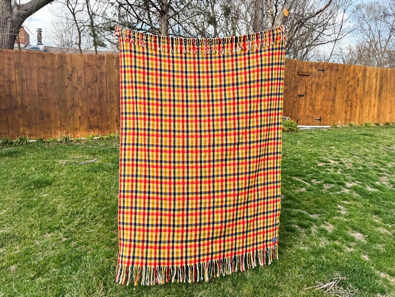 May include: A rectangular throw blanket featuring a vibrant plaid pattern in shades of yellow, red, and dark blue. The fabric has a woven texture and is finished with long, cream-colored fringe along the top and bottom edges of the textile.