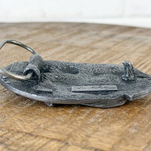 May include: A detailed, antique-style metal belt buckle with a silver-coloured loop and a textured, grey base. The buckle has a rectangular shape with a raised design and the name "Kent McSchering" is visible.