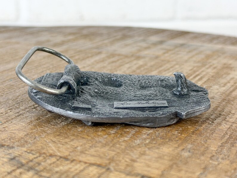 May include: A detailed, antique-style metal belt buckle with a silver-coloured loop and a textured, grey base. The buckle has a rectangular shape with a raised design and the name "Kent McSchering" is visible.