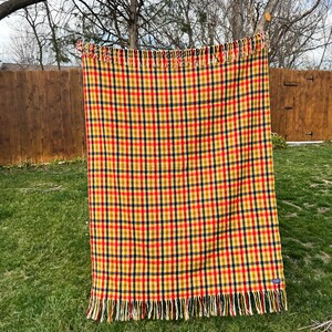 May include: A rectangular woven blanket featuring a vibrant plaid pattern in shades of red, yellow, and dark blue. The textile has a consistent grid design throughout and is finished with long, decorative fringe along the top and bottom edges.