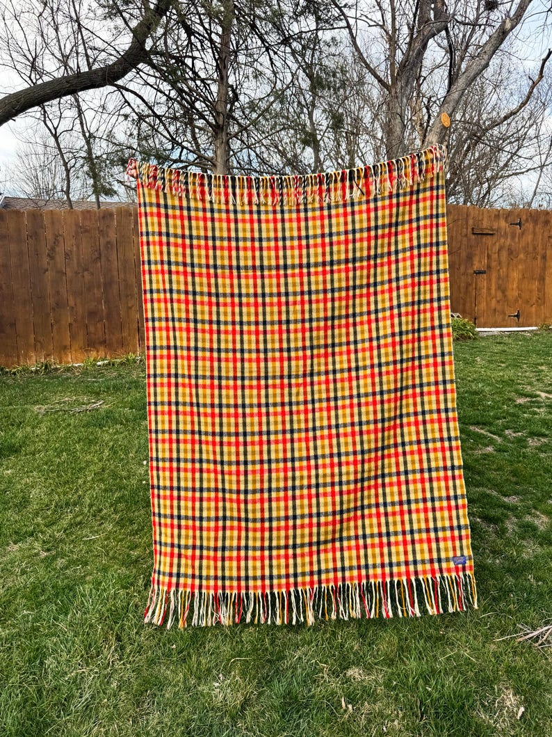 May include: A rectangular woven blanket featuring a vibrant plaid pattern in shades of red, yellow, and dark blue. The textile has a consistent grid design throughout and is finished with long, decorative fringe along the top and bottom edges.
