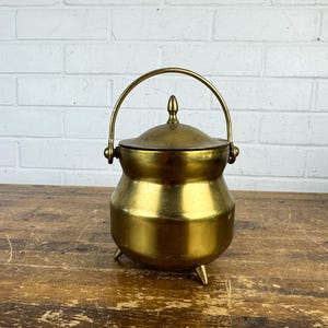 May include: A polished brass pot with a rounded body, three small feet, and a matching lid featuring a central finial. The vessel includes a curved, hinged handle attached to the sides, presenting a smooth metallic finish throughout the exterior.
