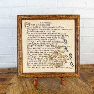 May include: A square cross-stitch embroidery in a brown wooden frame featuring the poem His Promise in black thread on beige fabric. The text is accompanied by a stitched illustration of a tree and a vertical trail of black footprints.