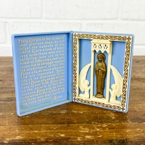 Pode incluir: Um ícone religioso azul articulado com uma estátua dourada de um santo. O lado esquerdo tem texto impresso. O lado direito tem uma moldura creme com asas de anjo. O texto diz "Holy Virgin...Amen."