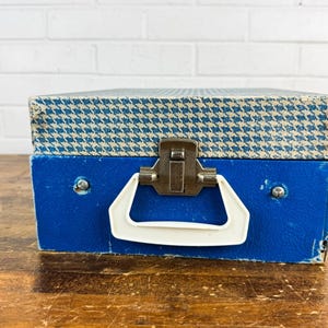 May include: A vintage blue and white houndstooth patterned box with a white handle and metal clasp. The box is a vibrant blue color and appears to be made of a sturdy material. The box is sitting on a wooden surface.