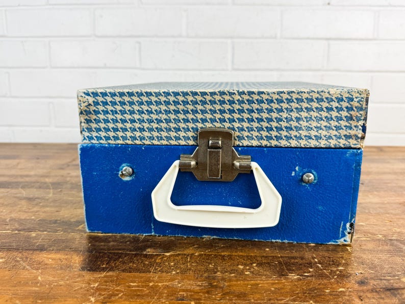 May include: A vintage blue and white houndstooth patterned box with a white handle and metal clasp. The box is a vibrant blue color and appears to be made of a sturdy material. The box is sitting on a wooden surface.