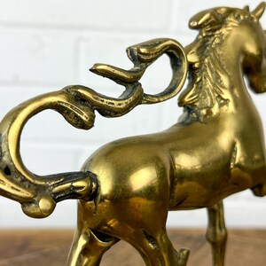 Puede incluir: Una figurita de unicornio de lat&oacute;n de color dorado con una cola rizada y un cuerno. El unicornio est&aacute; de pie sobre sus cuatro patas con la cabeza en alto.