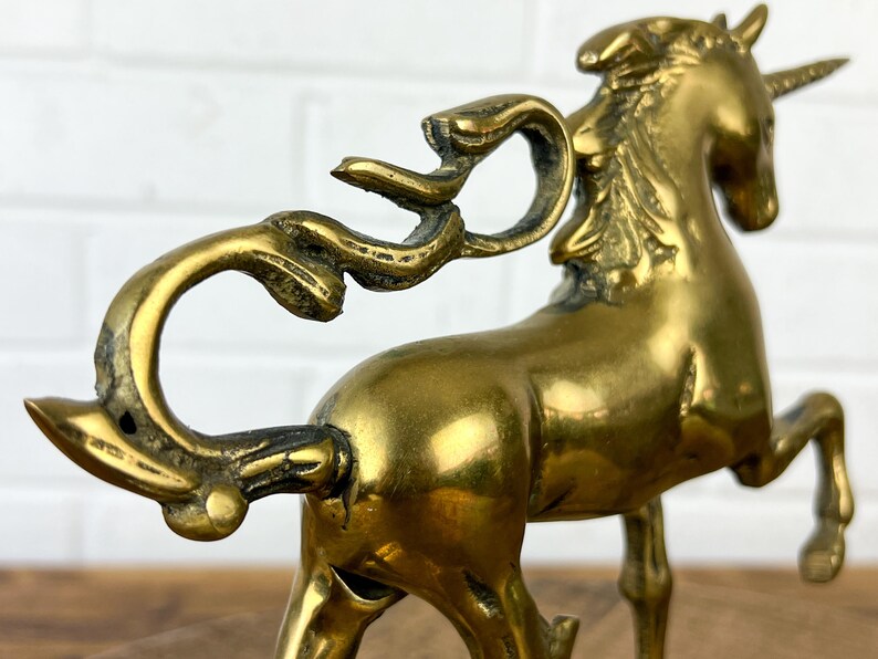 Puede incluir: Una figurita de unicornio de lat&oacute;n de color dorado con una cola rizada y un cuerno. El unicornio est&aacute; de pie sobre sus cuatro patas con la cabeza en alto.