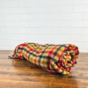 May include: A rolled-up wool blanket featuring a multi-colored plaid pattern in shades of mustard yellow, deep red, navy blue, and cream. The fabric has a textured weave and includes a fringed edge with loose threads at one end of the roll.