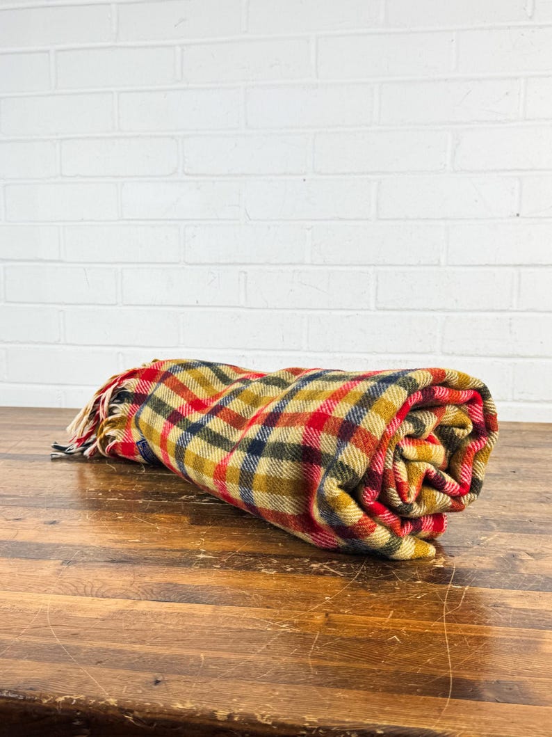 May include: A rolled-up wool blanket featuring a multi-colored plaid pattern in shades of mustard yellow, deep red, navy blue, and cream. The fabric has a textured weave and includes a fringed edge with loose threads at one end of the roll.