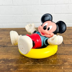 Puede incluir: Un juguete vintage de Mickey Mouse en un flotador amarillo. Mickey lleva pantalones cortos rojos, una camisa azul y guantes y zapatos blancos. El juguete est&aacute; sobre una superficie de madera con un fondo de ladrillos blancos.