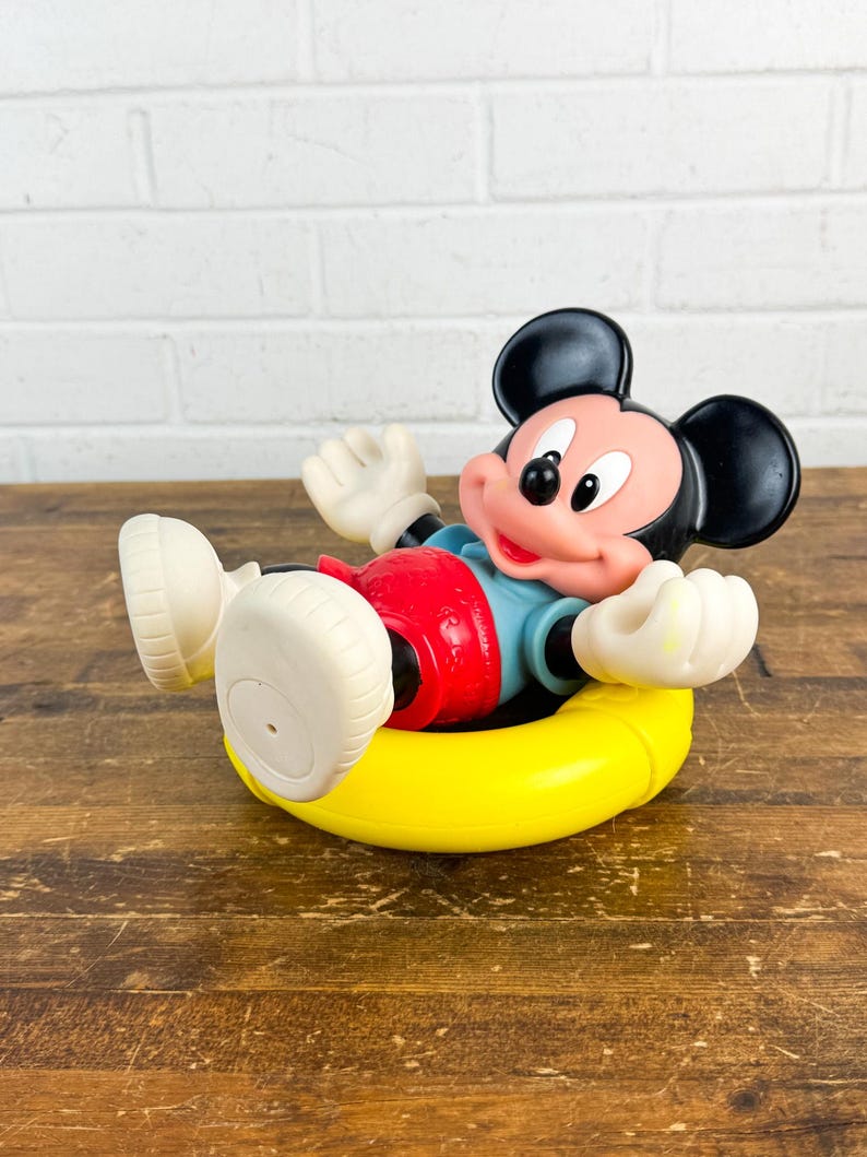 Puede incluir: Un juguete vintage de Mickey Mouse en un flotador amarillo. Mickey lleva pantalones cortos rojos, una camisa azul y guantes y zapatos blancos. El juguete est&aacute; sobre una superficie de madera con un fondo de ladrillos blancos.