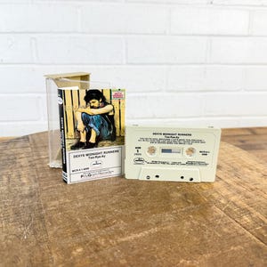 May include: A vintage cassette tape and case for the album "Too-Rye-Ay" by Dexys Midnight Runners. The cassette is off-white with black text, and the album cover features a man in a seated pose. The case is clear plastic.