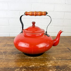 May include: A vintage red enamel teapot with a wooden handle and black metal accents. The teapot has a rounded body, a lid, and a curved spout. It sits on a wooden surface against a white brick wall.