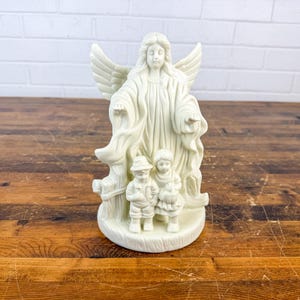 May include: A cream-colored angel figurine with outstretched wings and flowing robes. Two small children stand at the base of the angel. The figurine is made of a smooth material and is set against a wooden surface.
