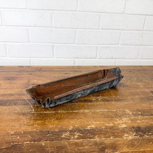 May include: An antique, rectangular metal trough with a weathered, rusty brown interior and dark gray exterior. The trough is set on a wooden surface with a white brick wall in the background. The trough has a folded, textured design.
