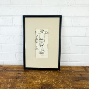 May include: A framed black and white line drawing on cream paper featuring two figures standing outdoors. The artwork is enclosed in a simple black frame with a wide cream-colored mat. The drawing depicts two individuals in casual attire.