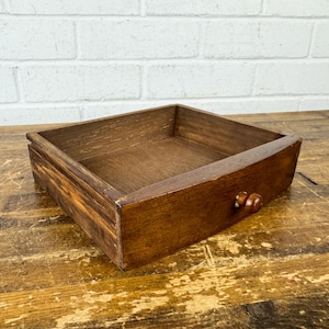 May include: A rectangular wooden drawer with a dark brown stained finish and a single small, round wooden knob centered on the front panel. The drawer features visible wood grain patterns and an open top, showcasing the interior construction.