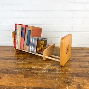 May include: A wooden book and CD holder with a wheelbarrow design. The holder is made of light-colored wood and holds several books and CDs. The books have red, blue, and tan covers. The CD case has the text "Offspring".