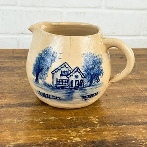 May include: A tan ceramic pitcher with a blue and white farmhouse design. The pitcher has a rounded body, a handle, and a spout. The farmhouse scene includes two houses, trees, and a fence. The pitcher is sitting on a wooden surface.