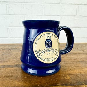 May include: A blue ceramic mug with a cream-colored owl logo. The logo reads "Wolfen's Roost 1915 Albany, NY".