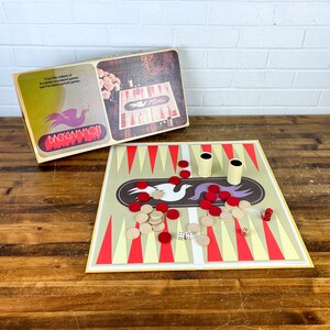 May include: A vintage backgammon game set with a brown and white game board, red and white playing pieces, and two dice. The game board has a design of two birds on it. The game is in a box with the word "Backgammon" on it.
