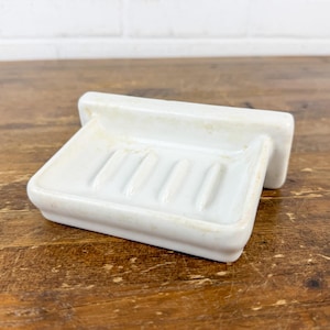 May include: A white ceramic soap dish with a rectangular base and a raised back. The dish has four parallel ridges to hold the soap and is set on a wooden surface.