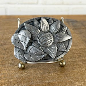 May include: A silver-coloured metal belt buckle with a detailed sunflower design and the word "KANSAS" in the centre. The oval-shaped buckle includes leaves. It is displayed on a small metal stand.