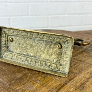 Puede incluir: Un sujetalibros de lat&oacute;n con un dise&ntilde;o decorativo. El sujetalibros tiene una base rectangular con un borde elevado y dos tornillos en la parte inferior.