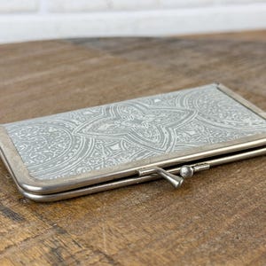 May include: A rectangular, silver-toned eyeglasses case with a decorative, light gray and white floral pattern. The case has a metal frame and a clasp closure. The case is closed and resting on a wooden surface.