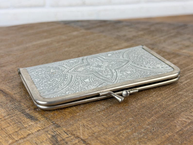 May include: A rectangular, silver-toned eyeglasses case with a decorative, light gray and white floral pattern. The case has a metal frame and a clasp closure. The case is closed and resting on a wooden surface.
