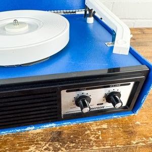 May include: A vintage blue Emerson record player with a white turntable and a black control panel. The control panel features tone and volume knobs. The record player sits on a wooden surface.