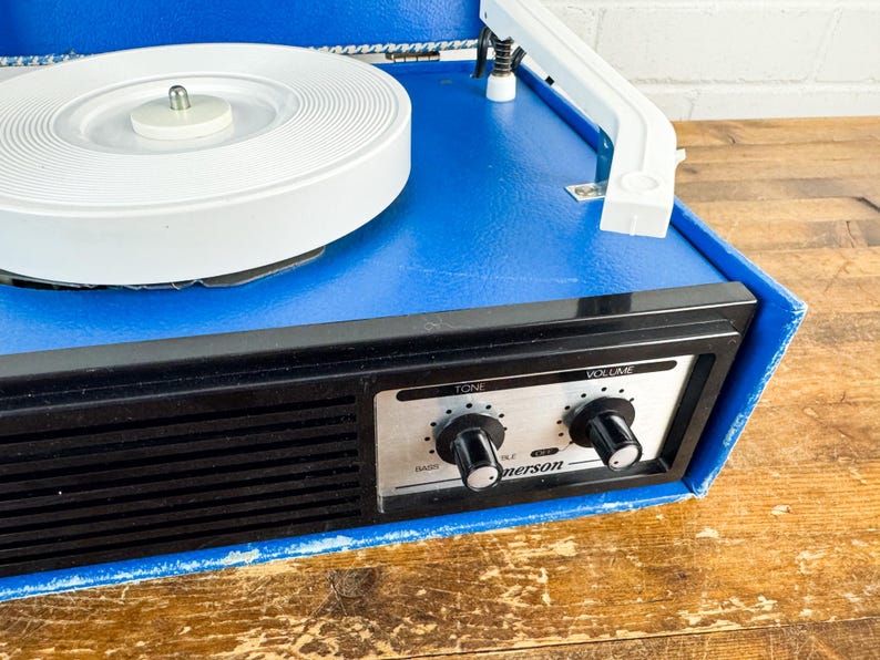 May include: A vintage blue Emerson record player with a white turntable and a black control panel. The control panel features tone and volume knobs. The record player sits on a wooden surface.