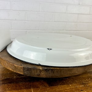 Puede incluir: Un plato de pastel de esmalte blanco con un borde negro se asienta sobre un pedestal de madera. El plato tiene algo de desgaste, lo que le da un aspecto vintage.