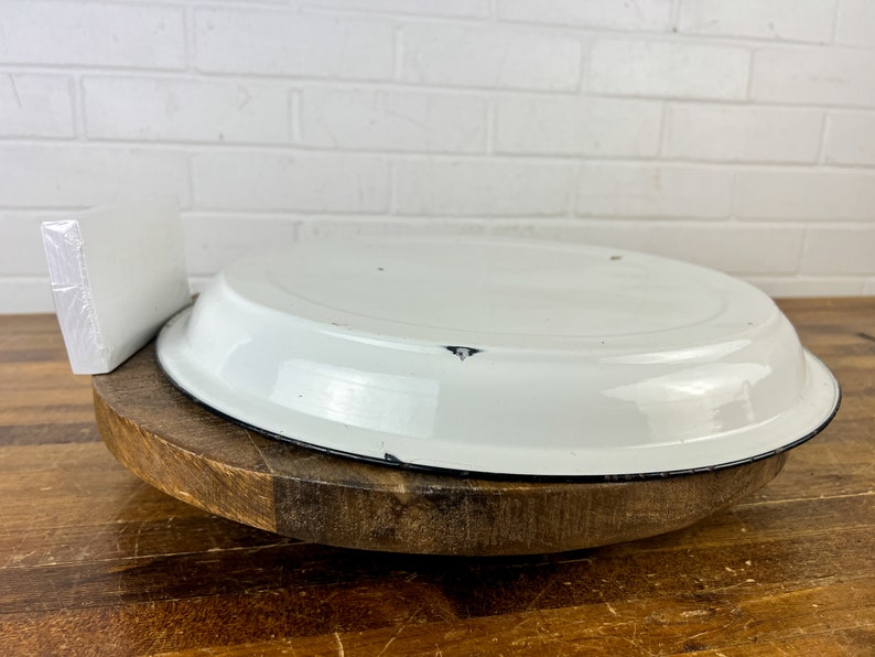 Puede incluir: Un plato de pastel de esmalte blanco con un borde negro se asienta sobre un pedestal de madera. El plato tiene algo de desgaste, lo que le da un aspecto vintage.
