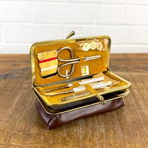 May include: A vintage brown leatherette sewing kit with a gold-toned metal frame. The kit is open, revealing various sewing tools, including scissors, needles, and buttons, set against a yellow velvet lining. The kit is on a wooden surface.