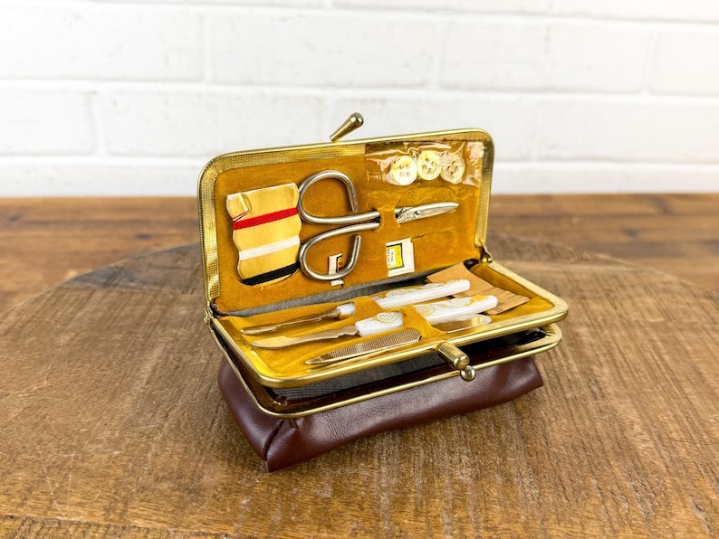 May include: A vintage brown leatherette sewing kit with a gold-toned metal frame. The kit is open, revealing various sewing tools, including scissors, needles, and buttons, set against a yellow velvet lining. The kit is on a wooden surface.