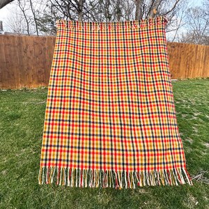 May include: A rectangular woven blanket featuring a multi-colored plaid pattern in shades of red, yellow, and dark blue. The fabric has a textured appearance and is finished with a fringe of loose threads along the top and bottom edges.