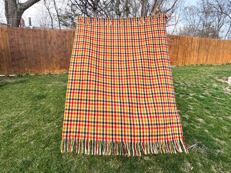 May include: A rectangular woven blanket featuring a multi-colored plaid pattern in shades of red, yellow, and dark blue. The fabric has a textured appearance and is finished with a fringe of loose threads along the top and bottom edges.