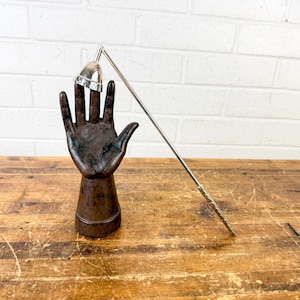 May include: A silver ring on a metal hand sculpture. The hand is dark brown and has a textured finish. A silver rod extends from the hand, holding a silver bell-shaped object. The background is a white brick wall and a wooden table.