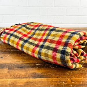 May include: A rolled-up woven blanket featuring a plaid pattern in shades of mustard yellow, bright red, and dark navy blue. The fabric has a textured appearance and includes a fringe detail on one end, consisting of loose, cream-colored threads.