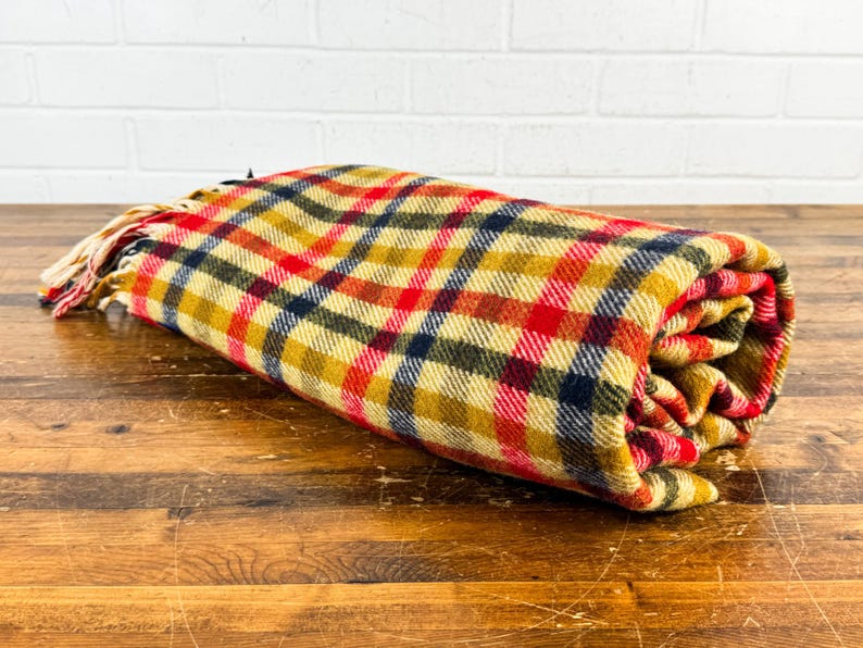 May include: A rolled-up woven blanket featuring a plaid pattern in shades of mustard yellow, bright red, and dark navy blue. The fabric has a textured appearance and includes a fringe detail on one end, consisting of loose, cream-colored threads.