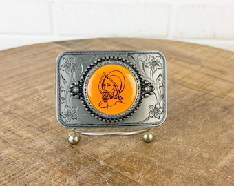 May include: A rectangular silver-toned metal belt buckle featuring an embossed floral border and a central circular inlay. The inlay has a bright orange background with a dark brown line drawing of a person wearing a historical helmet.