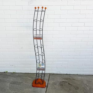 May include: A tall, freestanding, serpentine-shaped media storage rack constructed from thin black metal wire. The unit features four small orange spheres at the top and a matching orange base. It includes multiple horizontal slots for media cases.
