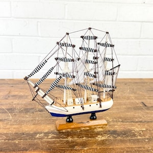 May include: A detailed wooden model of a sailing ship. The ship features white sails with black and white stripes, a white hull with a blue stripe, and intricate rigging. The model sits on a wooden base, showcasing nautical decor.