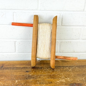 May include: A vintage wooden yarn swift with a spool of cream-colored yarn. The swift has two upright wooden supports with curved tops and orange handles. The yarn is tightly wound around the center.
