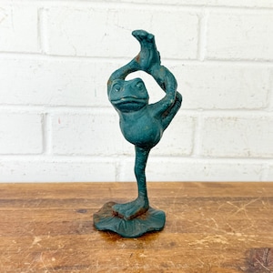 May include: A teal-colored cast iron frog sculpture in a yoga pose. The frog is standing on one leg, with the other leg and arm raised. The sculpture is on a brown wooden surface, with a white brick wall in the background.