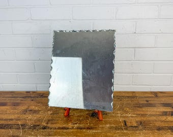 May include: A rectangular mirror with a scalloped, wavy edge design. The mirror is supported by a small, orange-coloured tripod stand. The reflective surface shows a clear, bright white reflection on the left side and a darker, muted tone on the right.