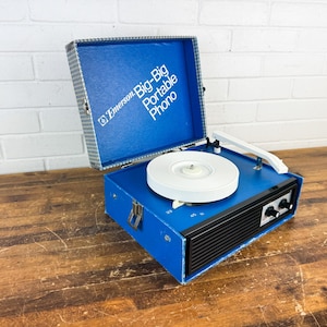 May include: A vintage Emerson Big-Big Portable Phono record player. The blue vinyl-covered case is open, revealing a white turntable and arm. The front has black speaker vents and control knobs. The case is on a wooden surface.